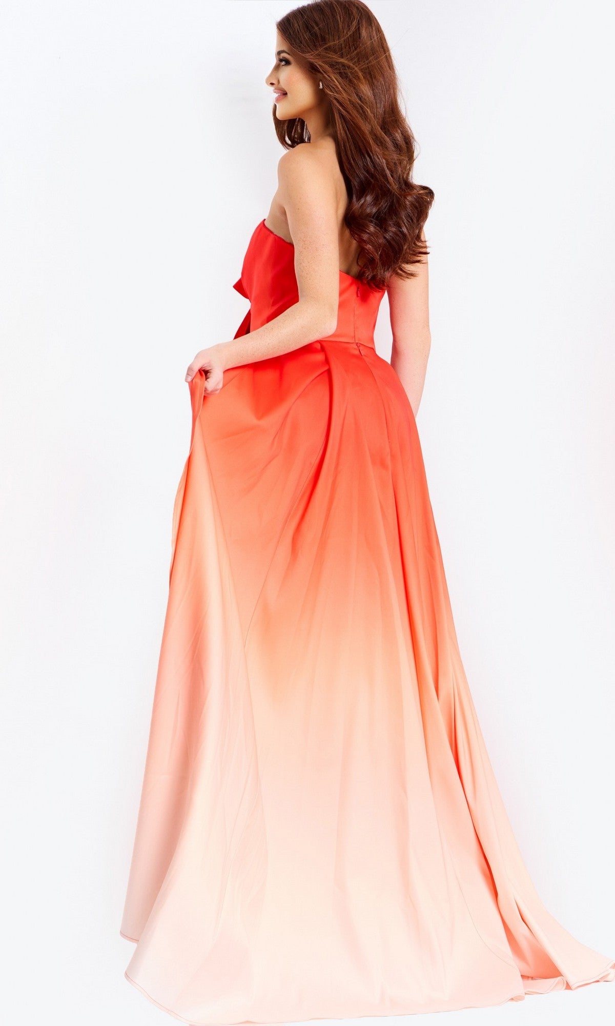 A woman wearing the Jovani 47554 long prom dress—an ombre strapless sheath style flowing from red to light peach—stands gracefully, holding part of the skirt.